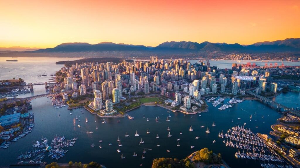 Aerial view of a waterfront city at sunset with a dense skyline, numerous sailboats in the harbor, and distant mountains on the horizon.