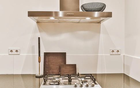 Stainless steel range hood above a gas cooktop in a modern kitchen, with white tiled backsplash, outlets on each side, and wooden cutting boards on the counter.